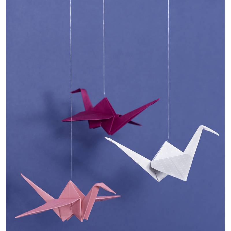 Drei Origami-Kraniche hängen an Schnüren vor einem lila Hintergrund, einer weiß, einer rosa und einer dunkelrosa, wodurch ein schwebender Effekt entsteht.