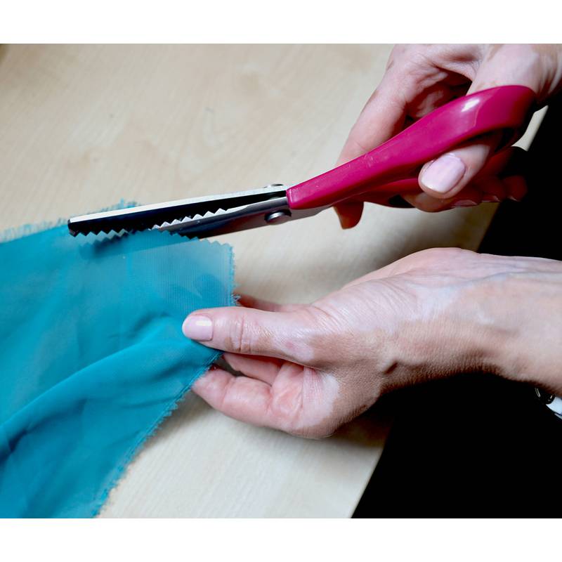 Eine Person schneidet blauen Stoff mit einer rosafarbenen Zickzackschere und demonstriert eine Technik zum Verhindern von Gewebekanten.