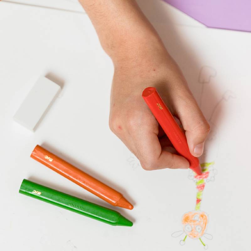 Eine Hand zeichnet eine farbenfrohe Figur auf weißem Papier mit einem pinken Wachsmalstift. In der Nähe liegen ein Radiergummi sowie grüne und orangene Wachsmalstifte auf dem Papier.