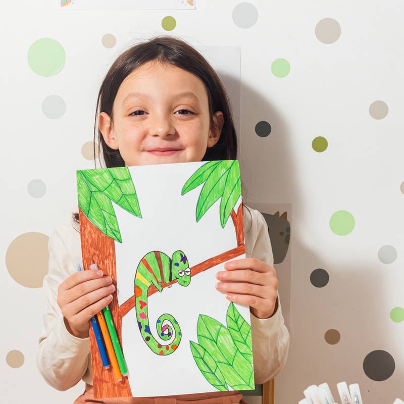 Ein Kind hält eine Zeichnung eines bunten Chamäleons auf einem Ast und lächelt stolz. Der Hintergrund zeigt Tapete mit Polkapunkten.
