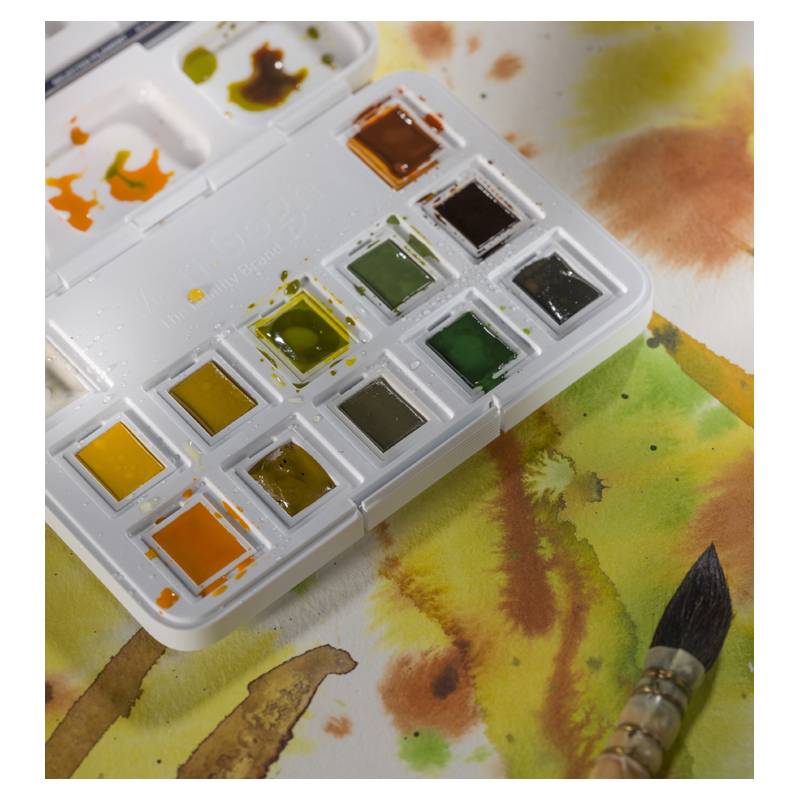 Eine offene Palette mit Aquarellfarben in verschiedenen Schattierungen von Grün, Gelb und Braun, neben einem Pinsel auf einer bemalten Oberfläche.