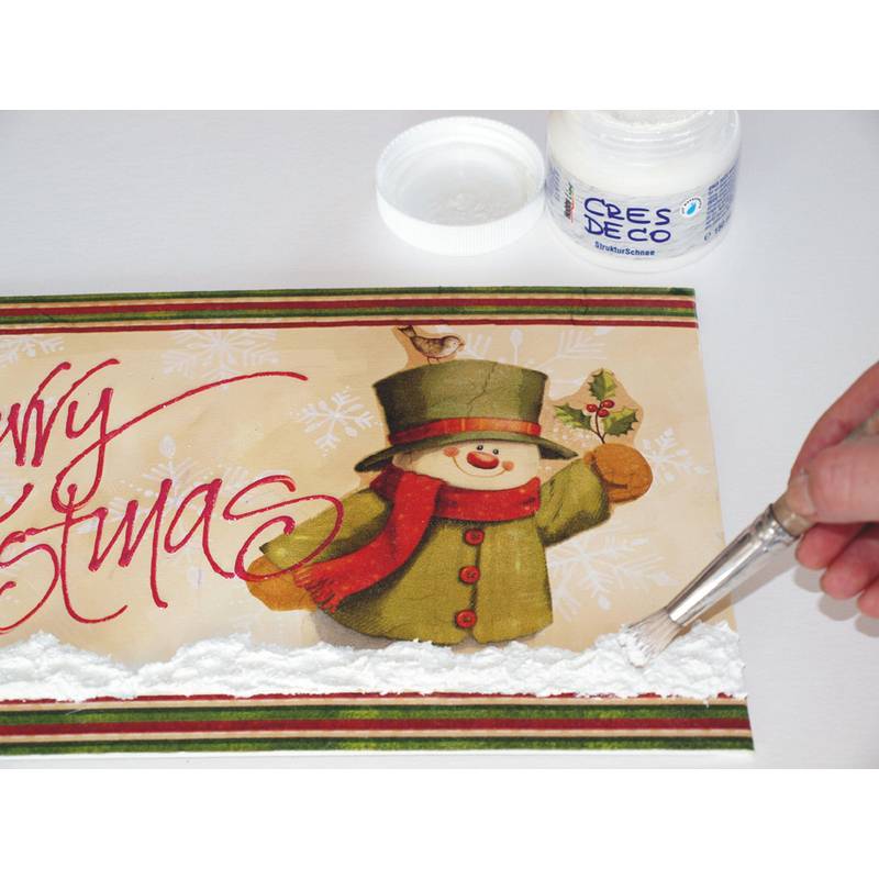 Eine Hand, die weiße strukturierte Paste aufträgt, um Schnee auf einer Weihnachtskarte zu gestalten, die einen Schneemann und den Text ‚Frohe Weihnachten' zeigt.