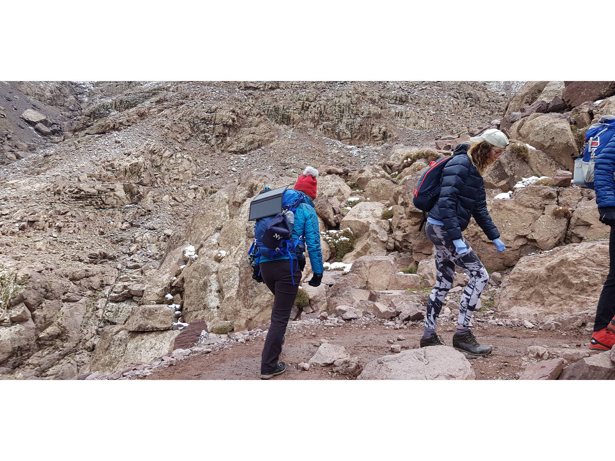 Drei Wanderer mit Rucksäcken erklimmen einen felsigen Bergpfad bei kaltem Wetter.