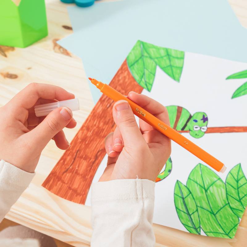 Eine Kinderhand zeichnet einen comicartigen Baum mit grünen Blättern und einem Ast auf Papier mit einem orangefarbenen Marker, vor einem Holztischhintergrund.