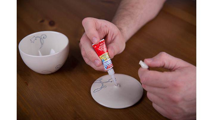 Hände tragen Klebstoff auf eine reparierte Keramikschale auf, neben ihr liegt eine Schüssel mit Bruchstücken.