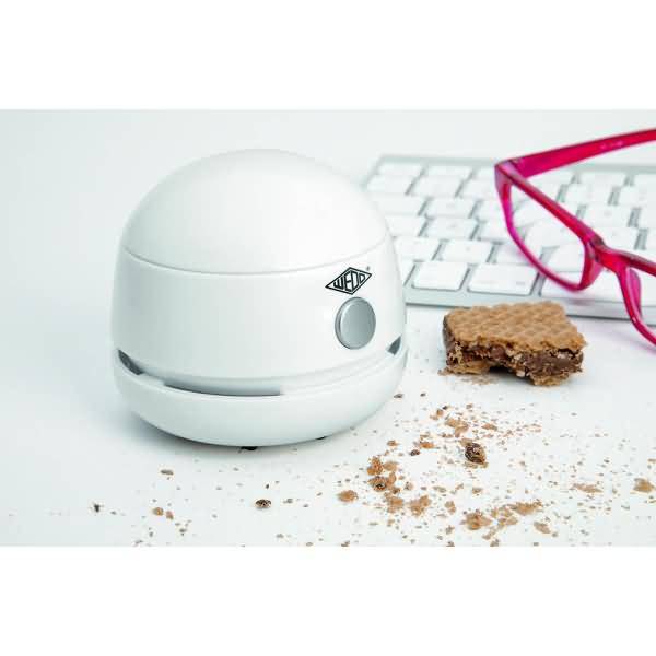 Tragbarer weißer Desktop-Staubsauger neben einer Tastatur und rosa Brille, mit Krümeln und einer halb aufgegessenen Waffel auf einer weißen Oberfläche.