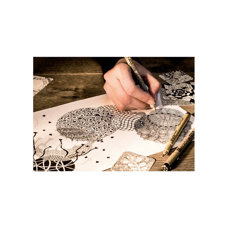 Eine Hand zeichnet komplizierte schwarz-weiße Muster auf Papier, wobei Stifte ringsum verteilt liegen und der Fokus auf detaillierten geometrischen Designs liegt.