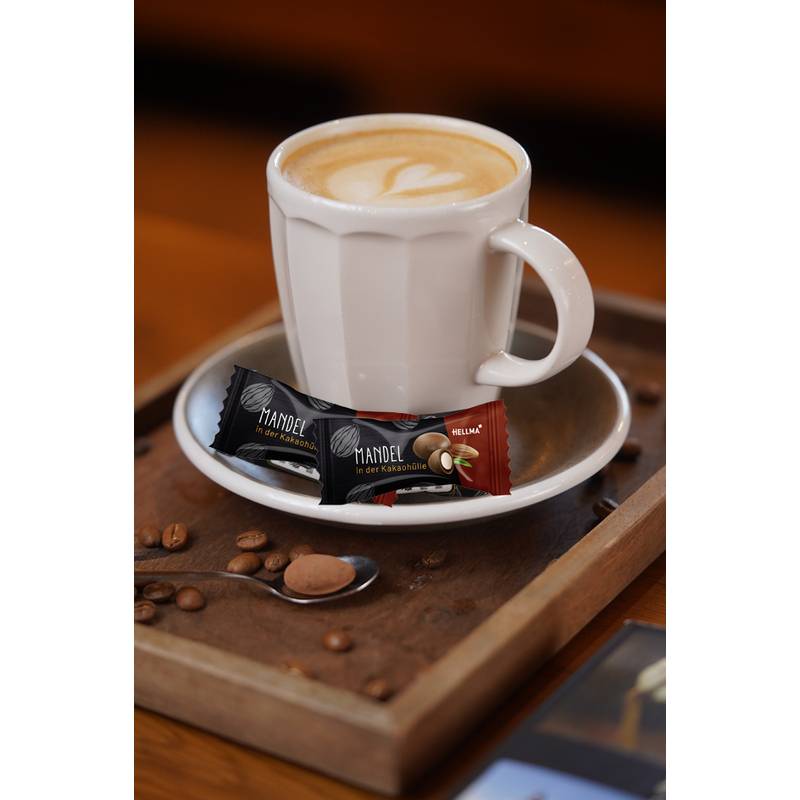 Eine Tasse Cappuccino auf einem Unterteller mit zwei verpackten Schokoladenbonbons daneben, platziert auf einem Holztablett mit Kaffeebohnen.