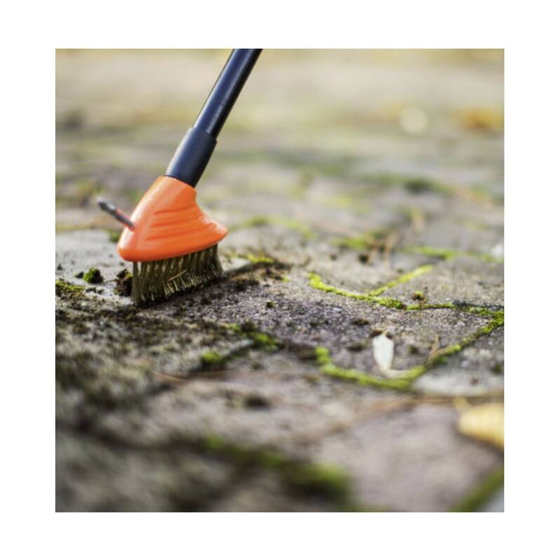 Ein Pinsel mit orangefarbenem Griff wird verwendet, um Moos und Schmutz zwischen Steinpflasterritzen zu reinigen, was auf eine Wartungs- oder Reinigungsaktivität hinweist.