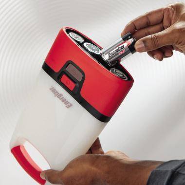 Hände führen zwei AA-Batterien in ein rot-weiß gefärbtes Energizer-Gerät ein, wahrscheinlich eine Taschenlampe oder tragbare Stromversorgungseinheit.