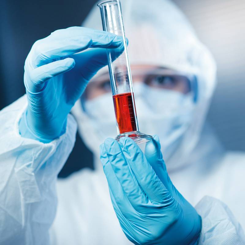 Ein Wissenschaftler in Schutzausrüstung hält ein Reagenzglas mit roter Flüssigkeit und untersucht es aufmerksam, was auf eine Laboranalyse oder wissenschaftliche Experimentaluntersuchung hindeutet.