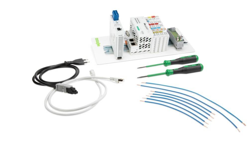 Eine weiße Grundplatte trägt eine Reihe elektronischer Komponenten, einschließlich eines weißen Moduls und Anschlussklemmen. Umgeben wird sie von einem schwarzen Kabel mit einem Netzstecker, einem weißen Netzwerkkabel, zwei Schraubenziehern mit grünen Griffen und einem Satz blauer Leitungen, in einer ordentlichen Anordnung.
