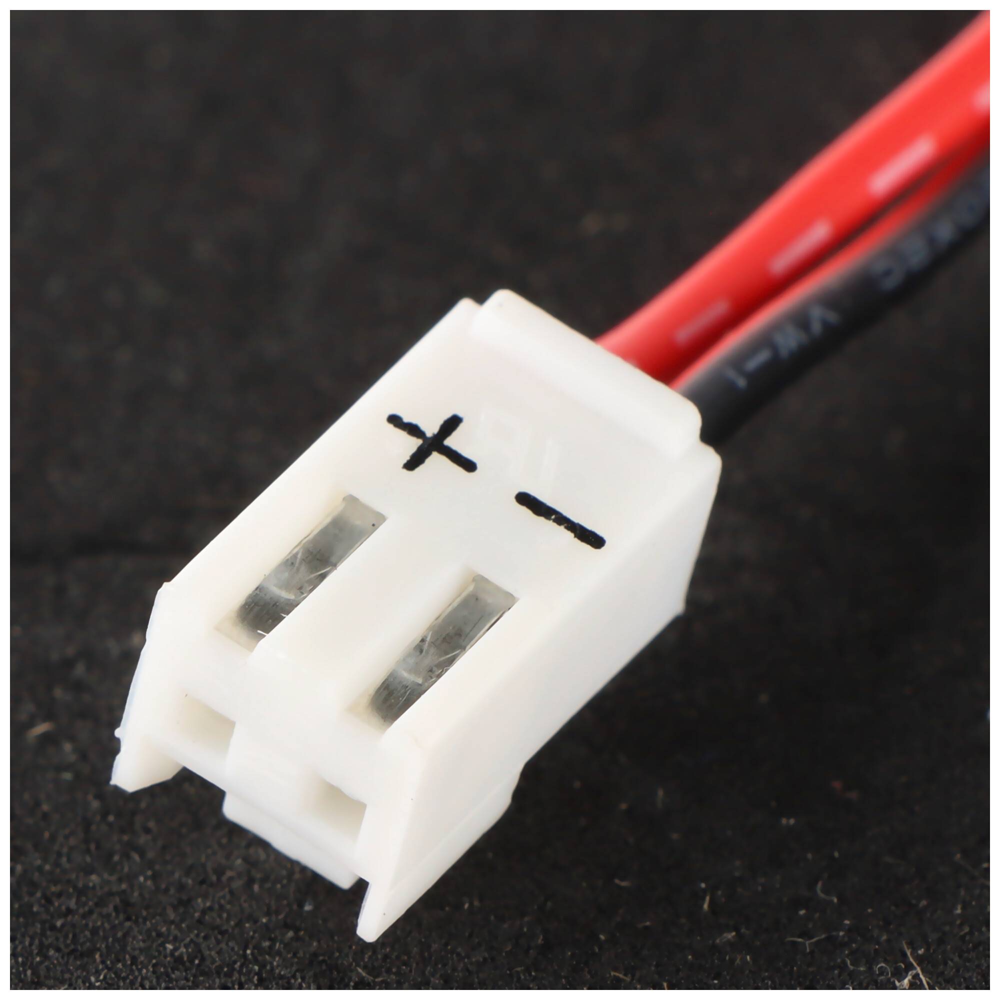 Nahaufnahme eines weißen zweiptigen Elektrosteckers mit roten und schwarzen Drähten, gekennzeichnet mit „+" und „-"", auf einer dunklen Oberfläche.