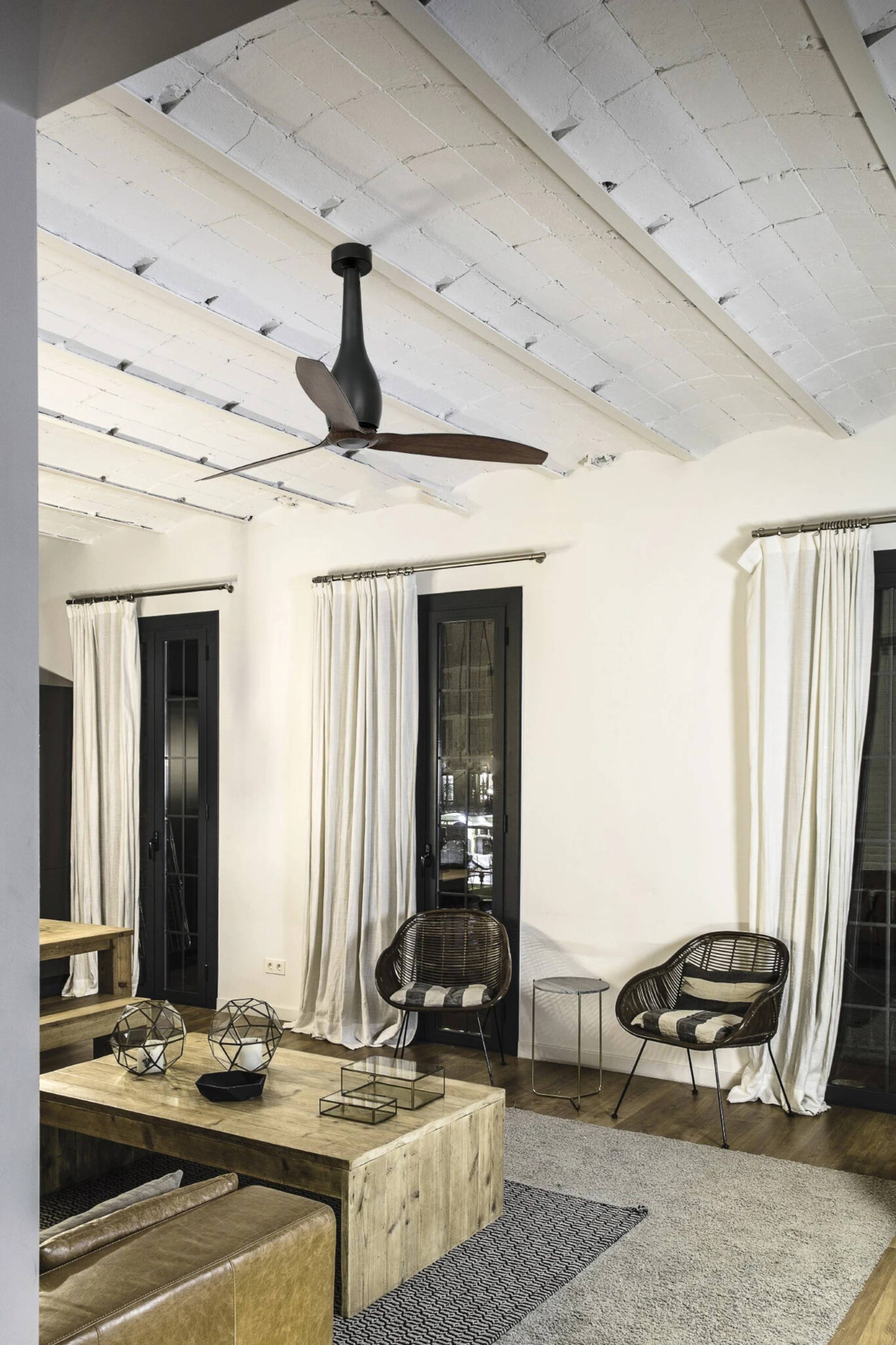 Ein modernes Wohnzimmer mit einem Deckenventilator, zwei Stühlen, einem Couchtisch und hohen Gardinen. Neutrale Töne dominieren die Einrichtung.