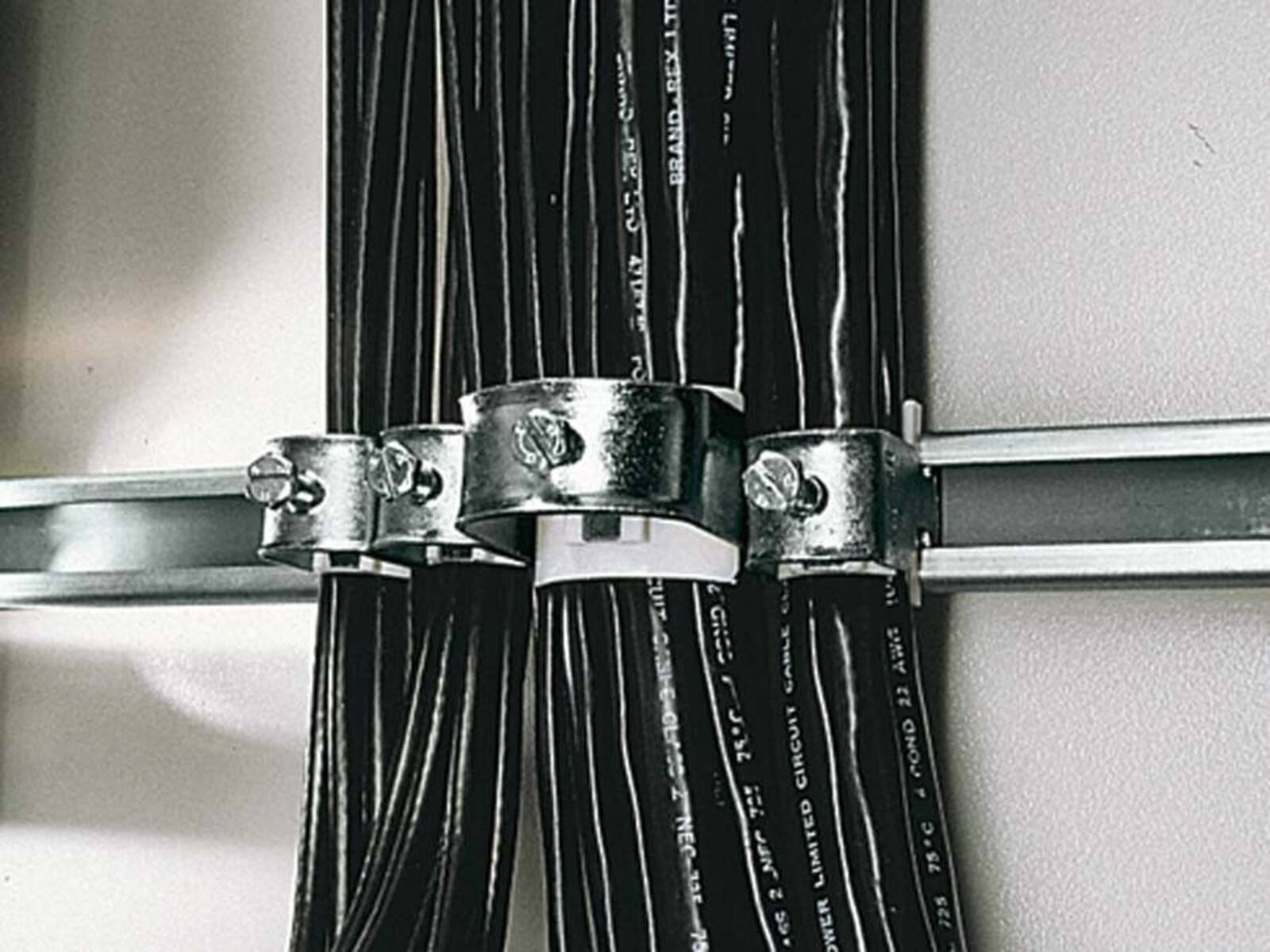 Schwarze Kabel, die mit Metallklemmen entlang einer Metallstange an einer Wand befestigt sind und eine organisierte und aufgeräumte Kabelführung vermitteln.