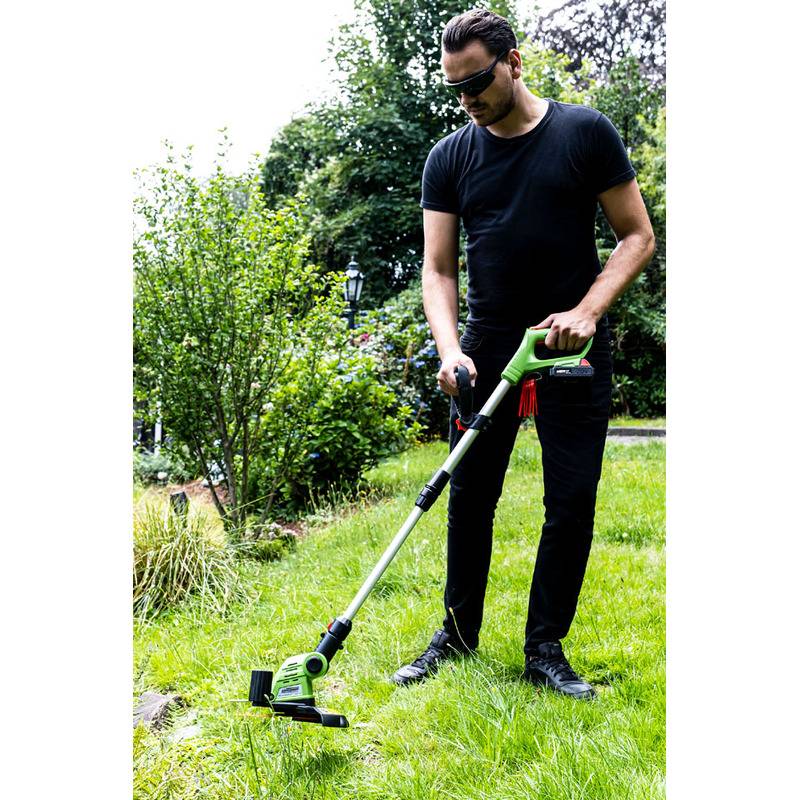 Eine Person mit Sonnenbrille benutzt einen grünen Trimmer, um Gras in einem Garten zu schneiden, umgeben von üppiger Vegetation und Bäumen im Hintergrund.