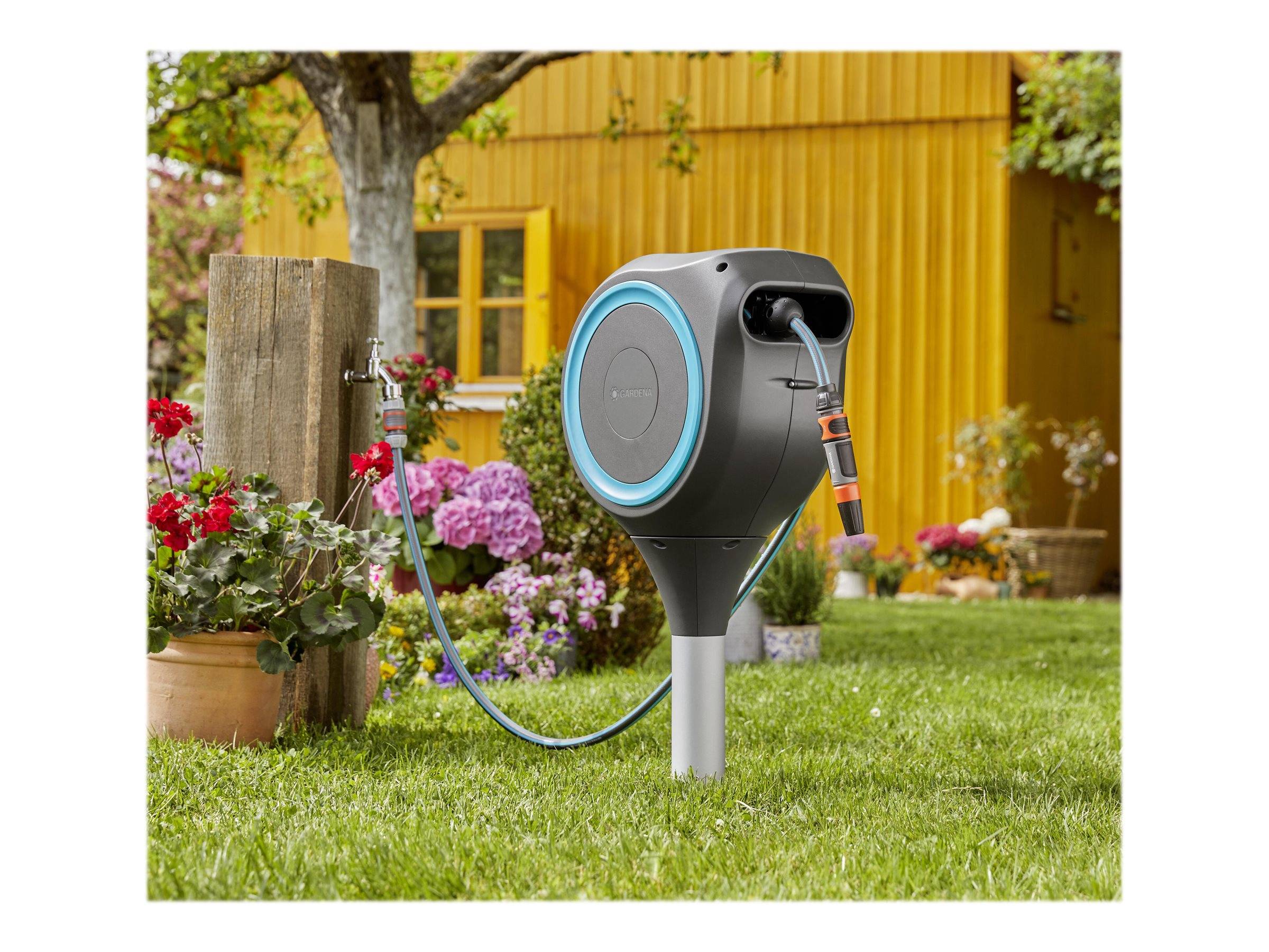 Tragbare Gartenschlauchtrommel auf einem Pfosten im Garten montiert, mit Schlauch verbunden mit einer Wasserquelle in der Nähe eines gelben Hauses und Blumen.