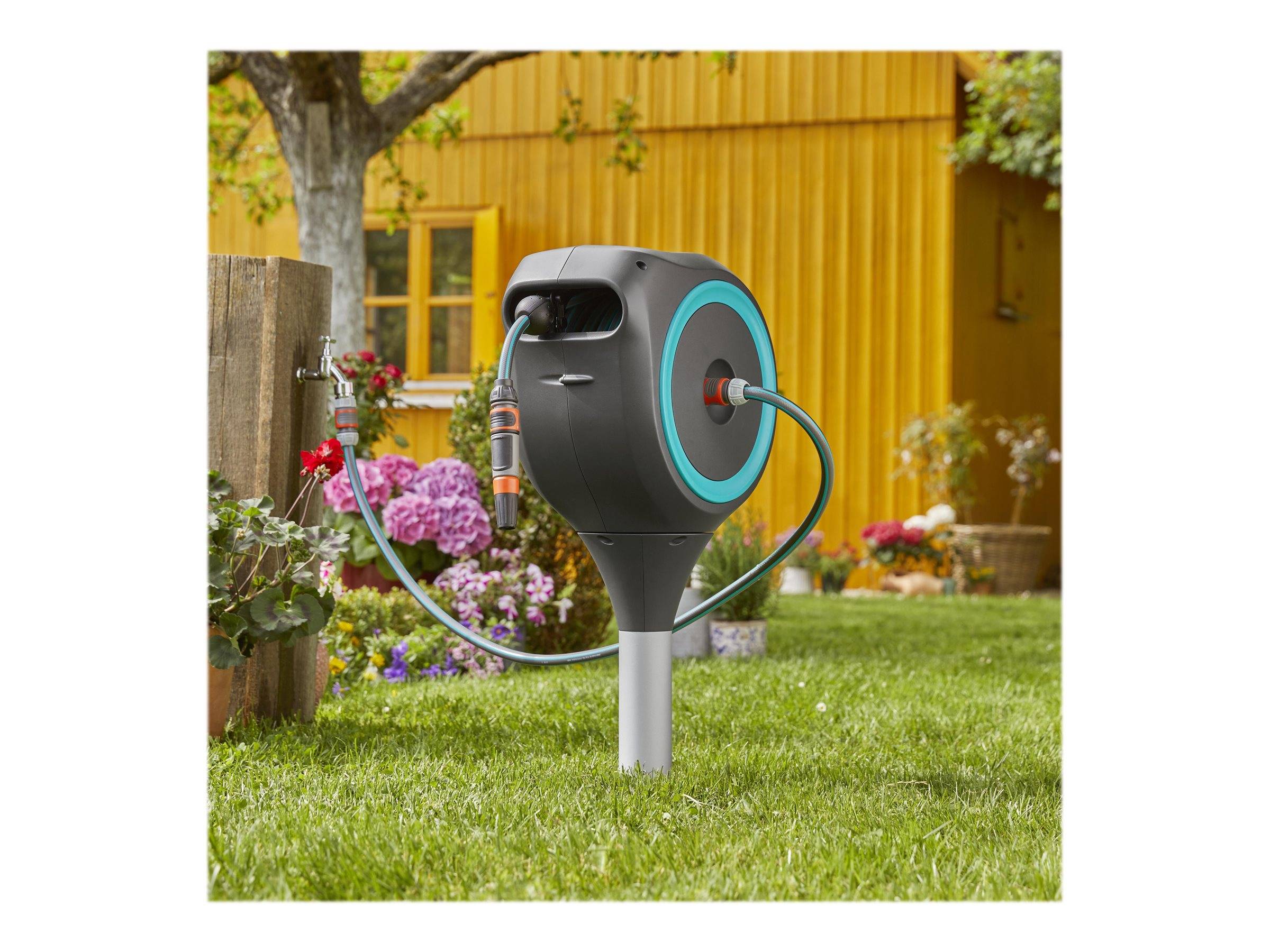 Eine Gartenschlauchrolle auf einem Metallgestell steht auf einem Rasen neben Blumen und einem gelben Haus, mit angebrachten und aufgewickelten Schläuchen.
