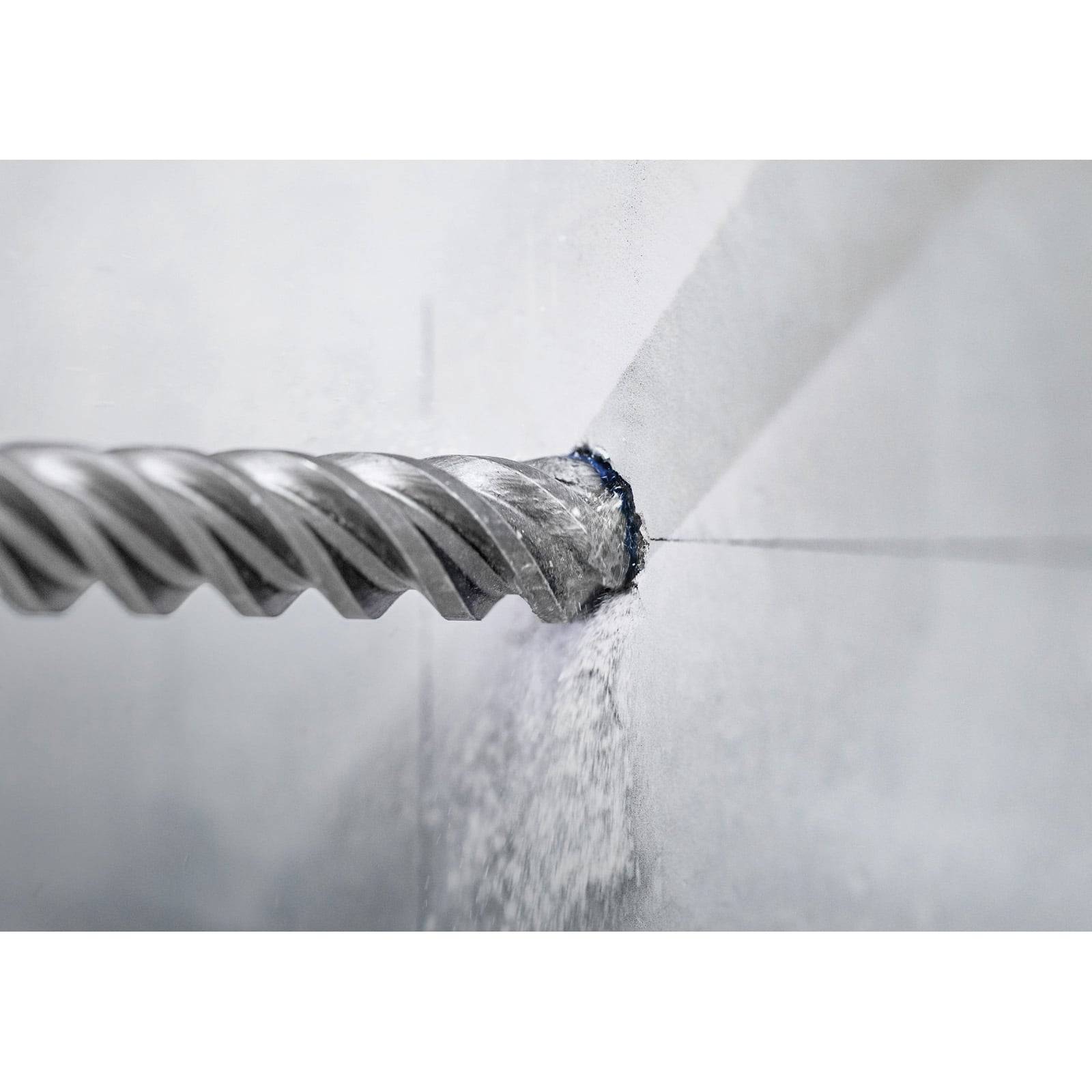 Eine Nahaufnahme eines Bohrers, der ein Loch in eine weiße Betonwand bohrt und dabei Staub erzeugt, was auf Bau- oder Renovierungsarbeiten hindeutet.