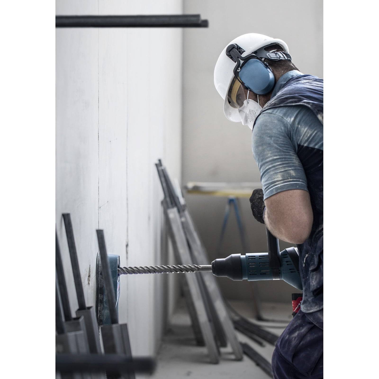 Ein Bauarbeiter mit Schutzhelm, Gehörschutz und Maske bohrt in einer unfertig wirkenden Raum in eine Wand und legt dabei Wert auf Sicherheit.