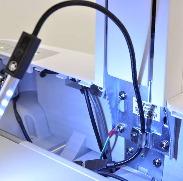Nahaufnahme des Inneren einer Industriemaschine mit einem flexiblen schwarzen Kabel, einer Metallhalterung, Bolzen und angebrachter Verdrahtung, beleuchtet durch blaues Licht.