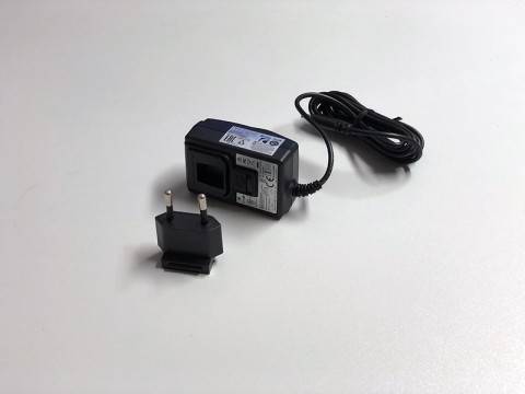 Schwarzer AC-Stromadapter mit angebundenem Kabel und abnehmbarem zweipoligem Stecker auf einer weißen Oberfläche.