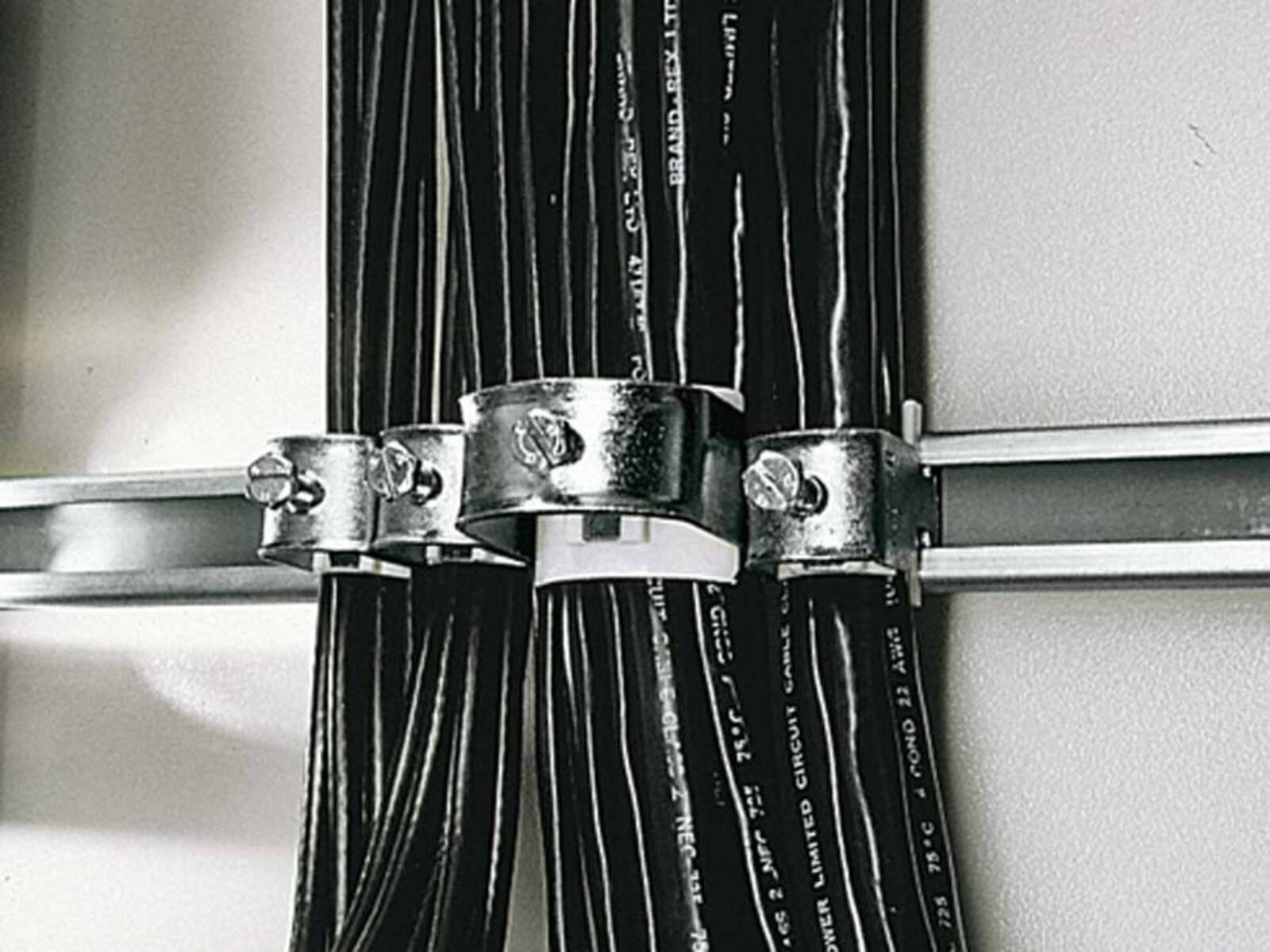Elektrisches Kabelbündel mit Metallklemmen an einer Wand befestigt, mit schwarzer Isolierung und weißen Textmarkierungen.