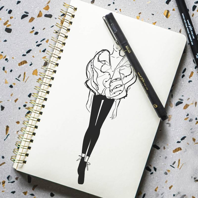 Eine Skizze eines Modedesigns mit einer weiblichen Figur in einem gerüschten Top und Hose, in schwarzer Tinte auf einem Notizbuch gezeichnet, mit einem schwarzen Stift darauf platziert.