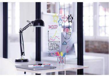Schreibtisch mit schwarzer Lampe, Laptop und transparenter Glastafeln, die bunte Geschäftskritzeleien, Diagramme und das Wort "ERFOLG"" in einer Büroumgebung zeigen