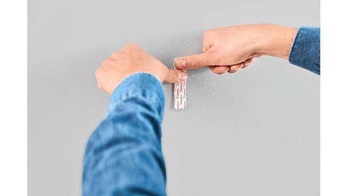 Hände schälen ein kleines Haftpflaster von einem Unterarm einer Person gegen einen einfachen grauen Hintergrund ab.