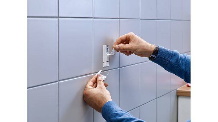 Hände schälen Schutzfolie von einem wandmontierten Haken oder Klebestreifen auf einer gefliesten Badezimmerwand ab und installieren ihn.