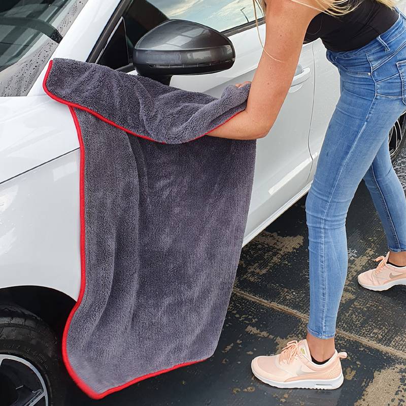 Eine Person, die ein weißes Auto mit einem grauen Handtuch reinigt und sich dabei auf den Außenspiegel der Fahrerseite konzentriert. Die Person trägt blaue Jeans und pinke Schuhe.