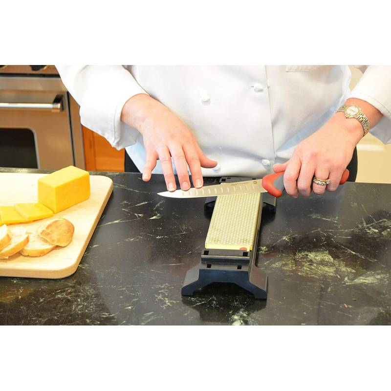 Eine Person, die ein großes Küchenmesser auf einem Wetzstein an der Küchenarbeitsplatte schärft, mit Käse und geschnittenem Brot auf einem nahegelegenen Holzbrett.