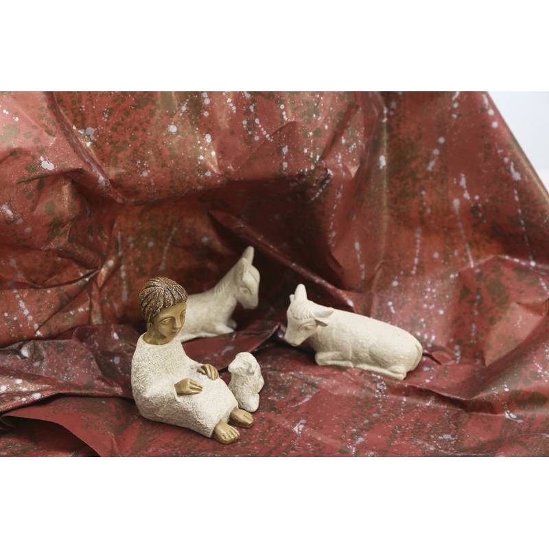 Eine kleine Figur einer Person in einem Gewand, sitzend mit einem Lamm zu ihren Füßen. In der Nähe liegen ein Esel und eine Kuh auf einer strukturierten roten Oberfläche.