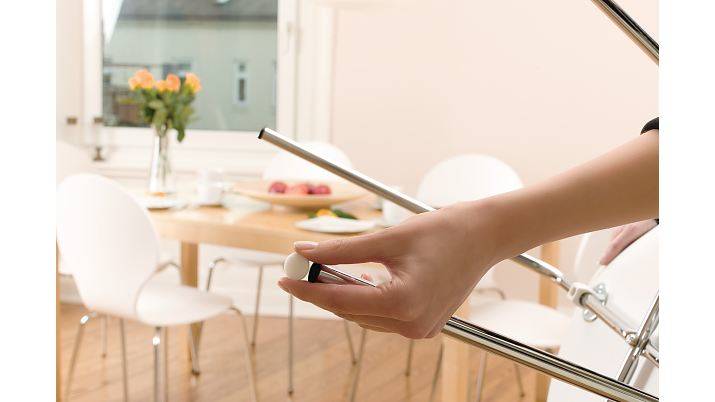 Eine Hand, die eine verchromte Metallstange in einem hellen Esszimmer mit Tisch, Stühlen, Blumen und Obst im Hintergrund hält.