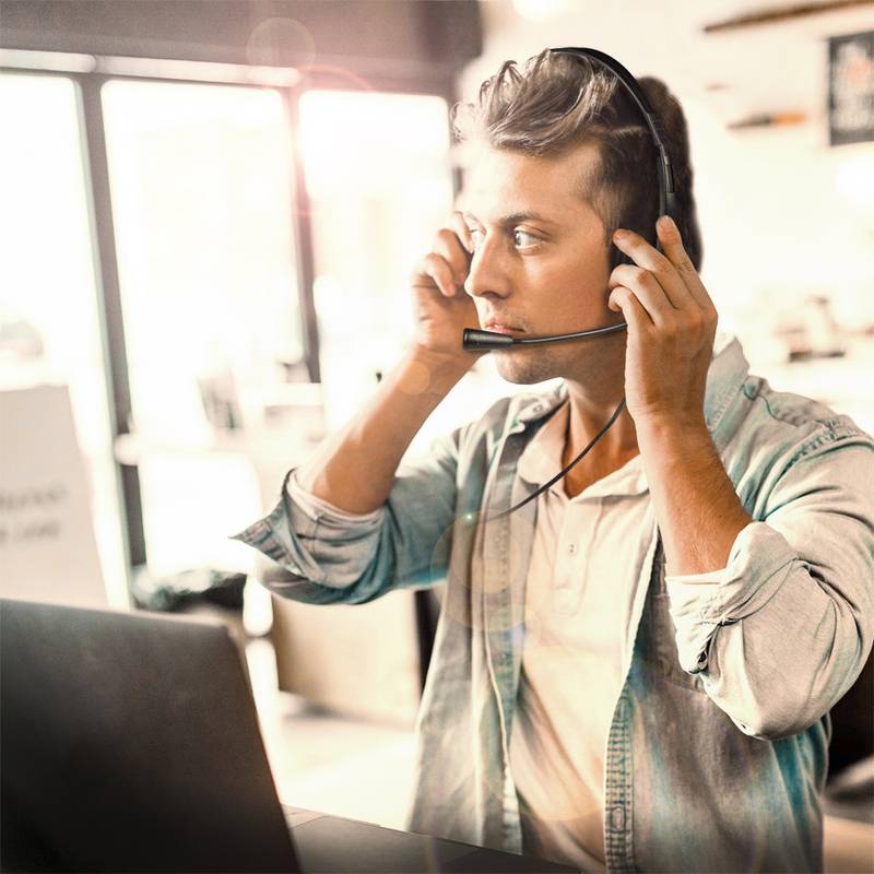 Eine Person mit einem Headset sitzt an einem Schreibtisch mit einem Laptop und konzentriert sich auf ein Gespräch in einer hell beleuchteten Büroumgebung.