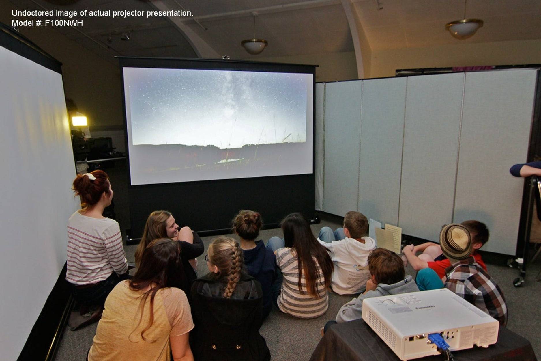 Kinder und Erwachsene sitzen auf dem Boden und schauen sich ein projiziertes Bild in einem dunklen Raum an, wobei ein Projektor im Vordergrund zu sehen ist.