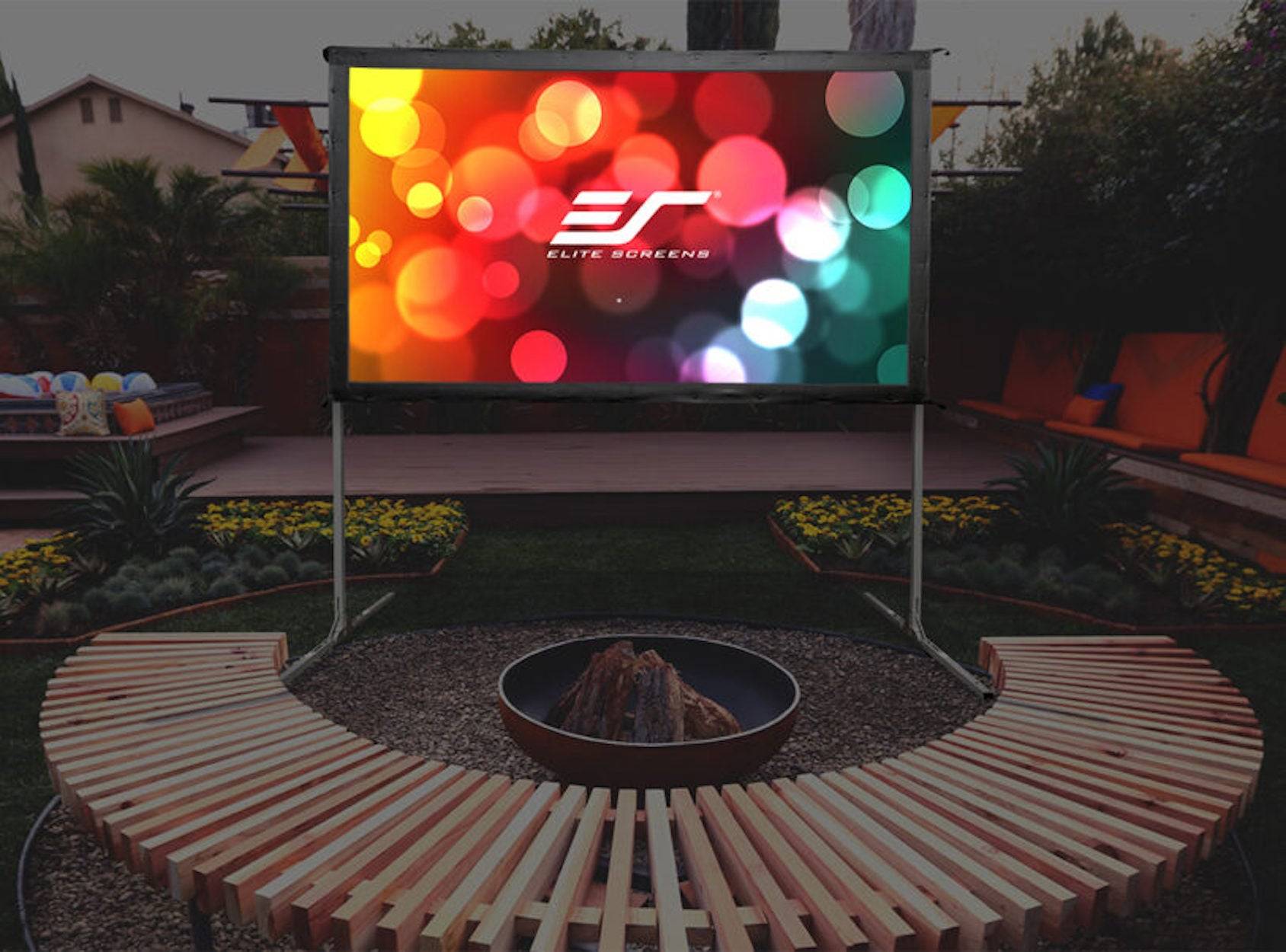 Outdoor-Projektionsleinwand im Hinterhof zeigt das Logo von "Elite Screens"", angebracht über einer geschwungenen Holzbank und einer Feuerstelle in der Dämmerung.