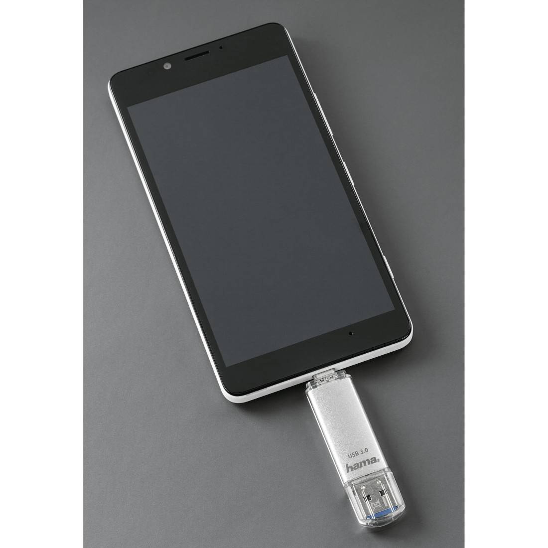 Schwarzes Smartphone, das mit einem silbernen USB-Speicherstick auf grauem Hintergrund verbunden ist und eine Telefon-zu-Speicher-Übertragung oder Sicherung zeigt.