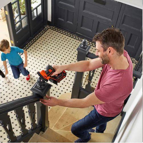 Ein Mann installiert mit einer kabellosen Bohrmaschine eine schwarze Handlaufhalterung auf einer Treppe, während ein Kind in der Nähe in einem Flur eines Hauses steht.