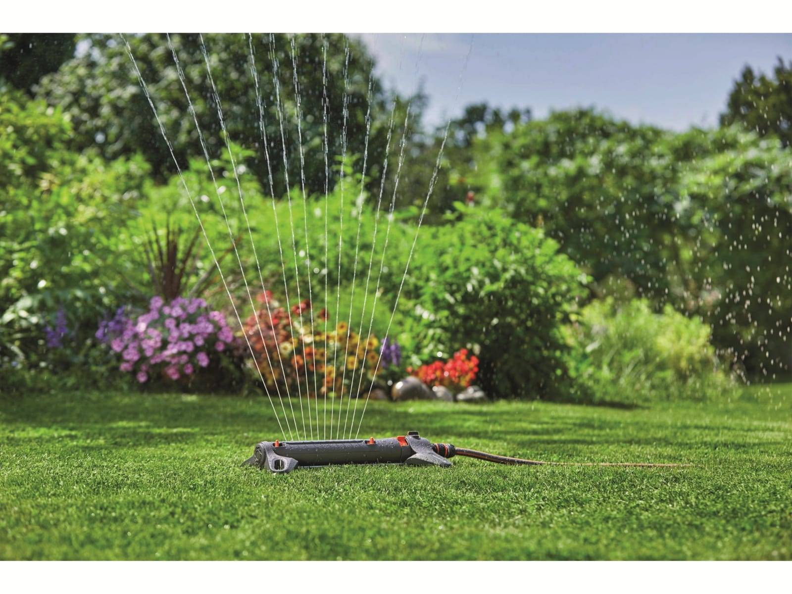 Oszillierender Rasensprenger bewässert einen üppig grünen Garten mit blühenden Sträuchern im Hintergrund.