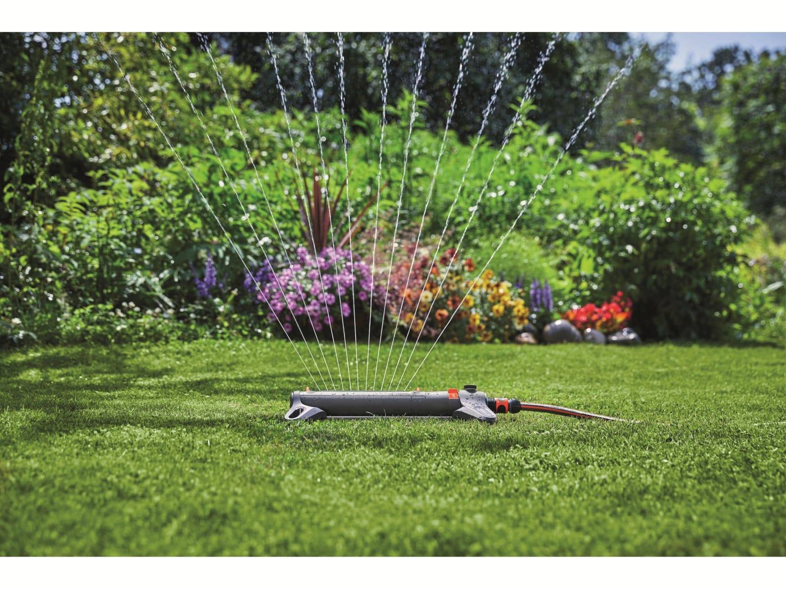 Ein Rasensprenger bewässert einen grünen Garten mit Blumen und Sträuchern im Hintergrund.