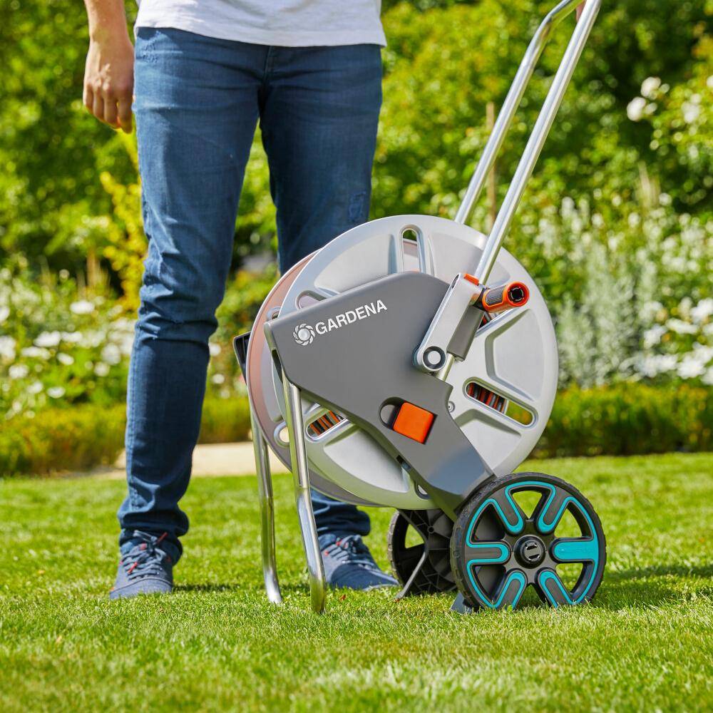 Person, die neben einer GARDENA-Schlauchrolle auf einem Rasen steht, mit einem fahrbaren Gartenschlauch-Wagen und metallischem Griff in einem grasbewachsenen Hof.