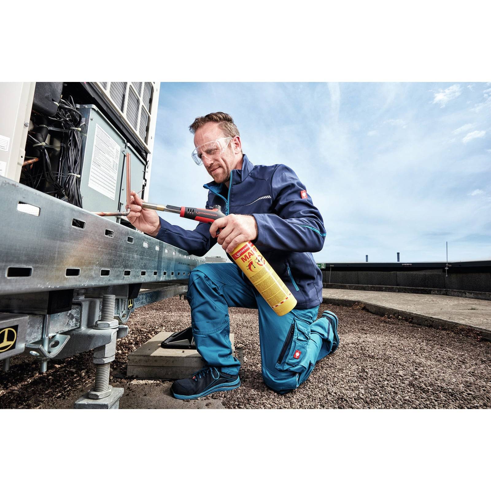 Techniker kniet neben einer Dachklimanlage und verwendet ein Lötgerät und einen gelben Gasbehälter, um im Freien an der Ausrüstung zu arbeiten.