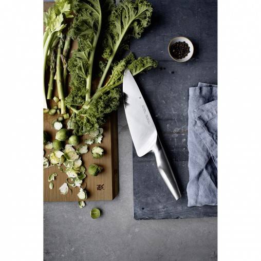 Frisches Grünkohl, Rosenkohl und ein Kochmesser auf einem Schneidebrett mit Pfefferkörnern und einem grauen Tuch auf einer dunklen Arbeitsplatte.