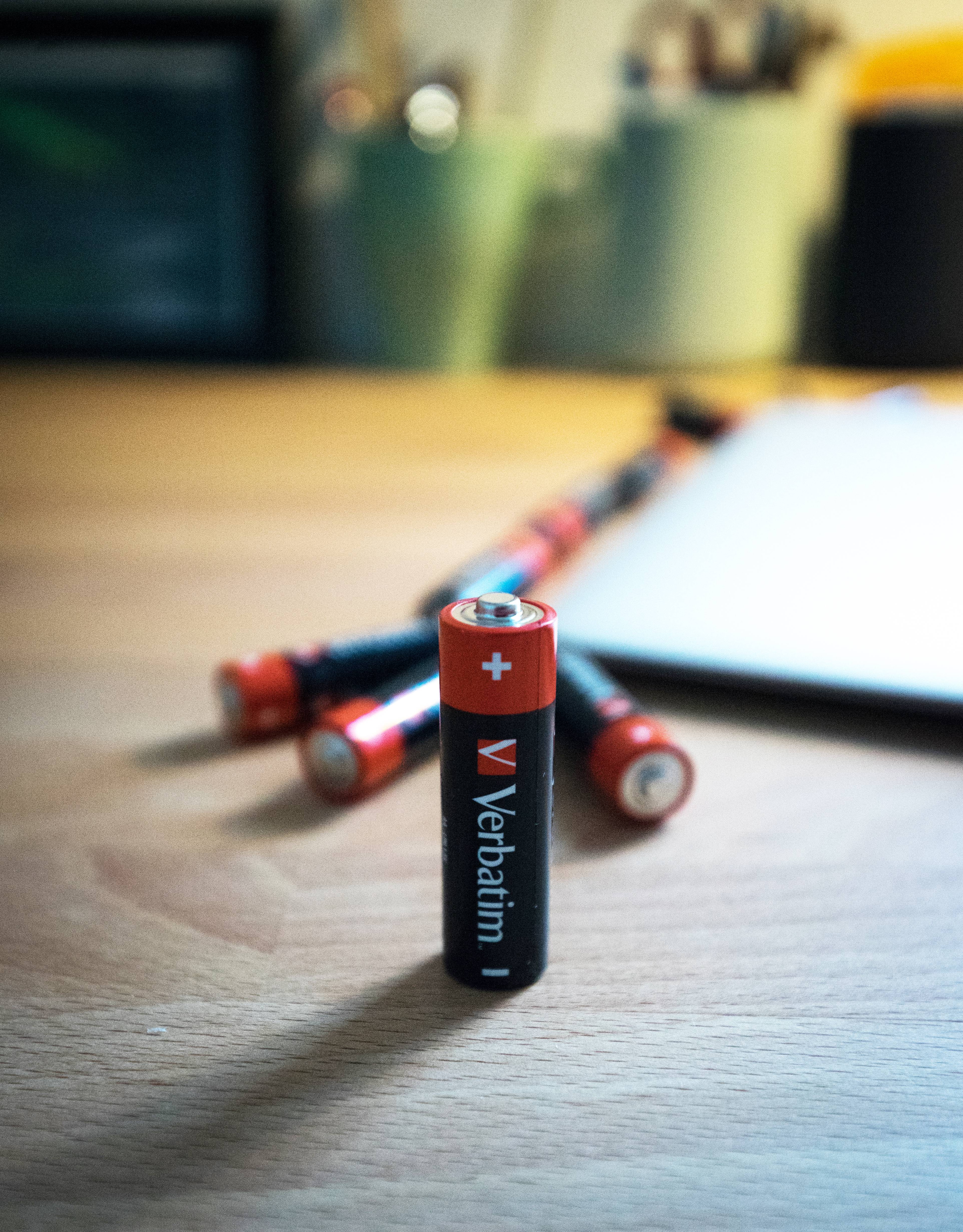 Eine aufrecht stehende Verbatim AA-Batterie auf einem Schreibtisch, mit mehreren anderen Batterien, die dahinter in der Nähe eines Notizblocks verstreut sind.