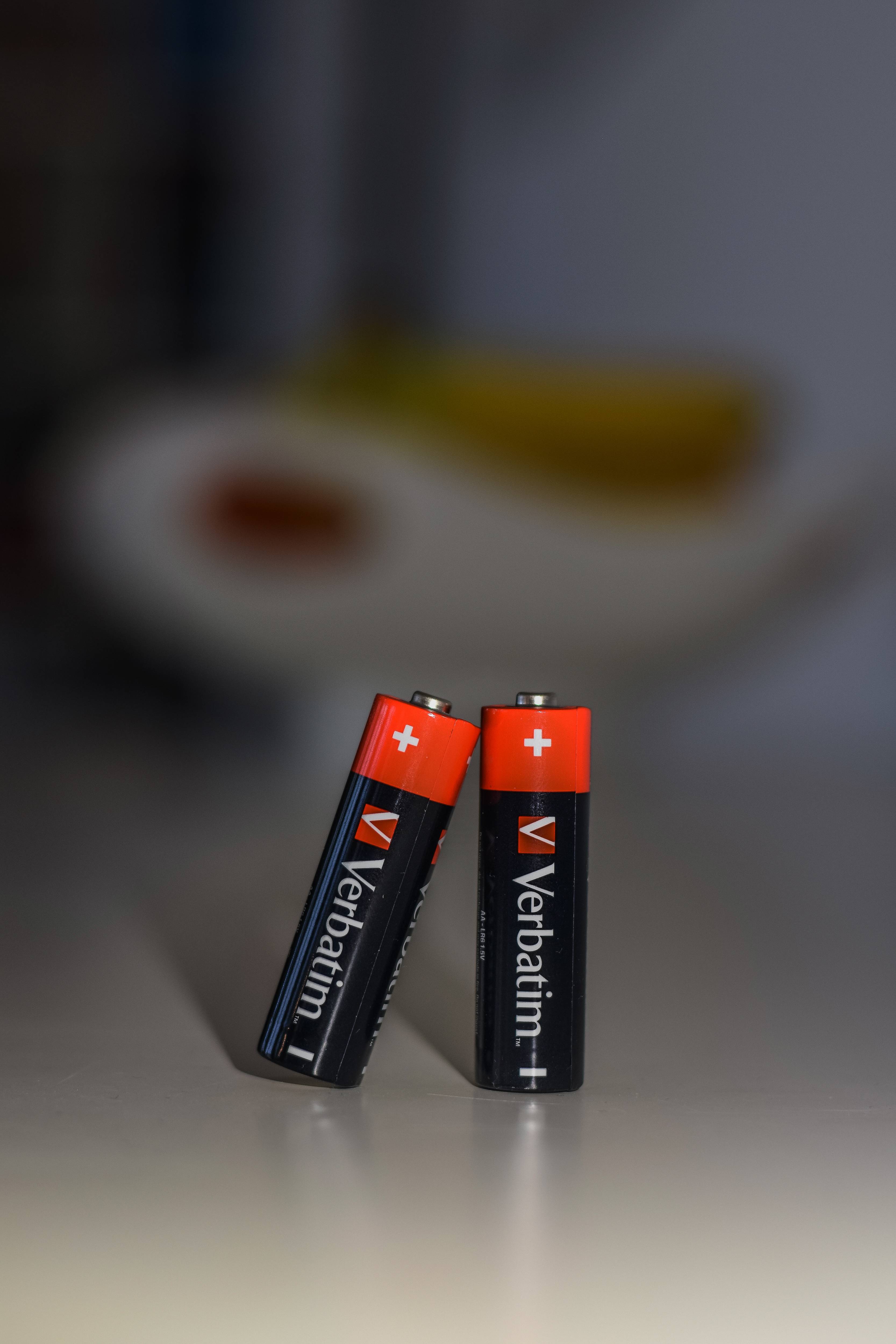 Zwei aufrecht stehende Verbatim-AA-Batterien auf einer reflektierenden Oberfläche, mit unscharfem Hintergrund.