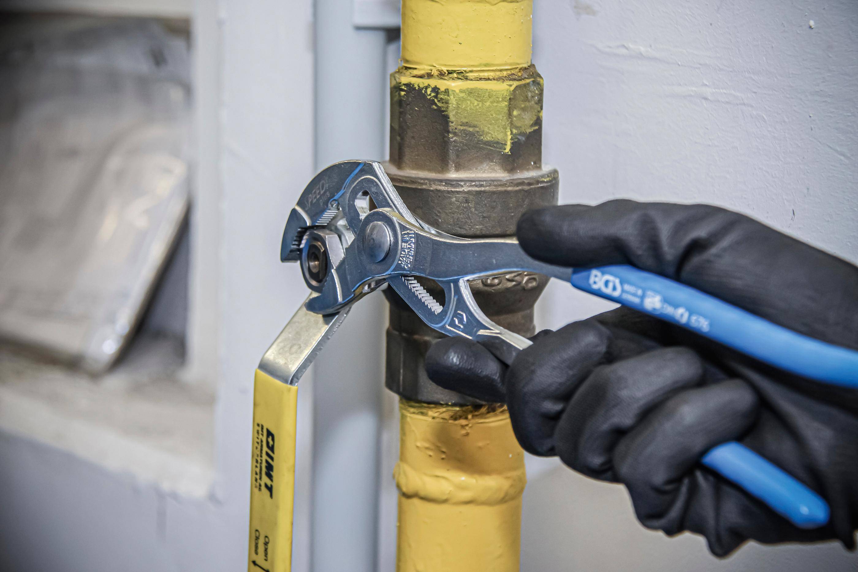 Eine behandschuhte Hand verwendet einen Schraubenschlüssel, um eine Verbindung an einem gelb lackierten Rohr festzuziehen oder zu lockern, was eine Wartungs- oder Reparaturaufgabe veranschaulicht.