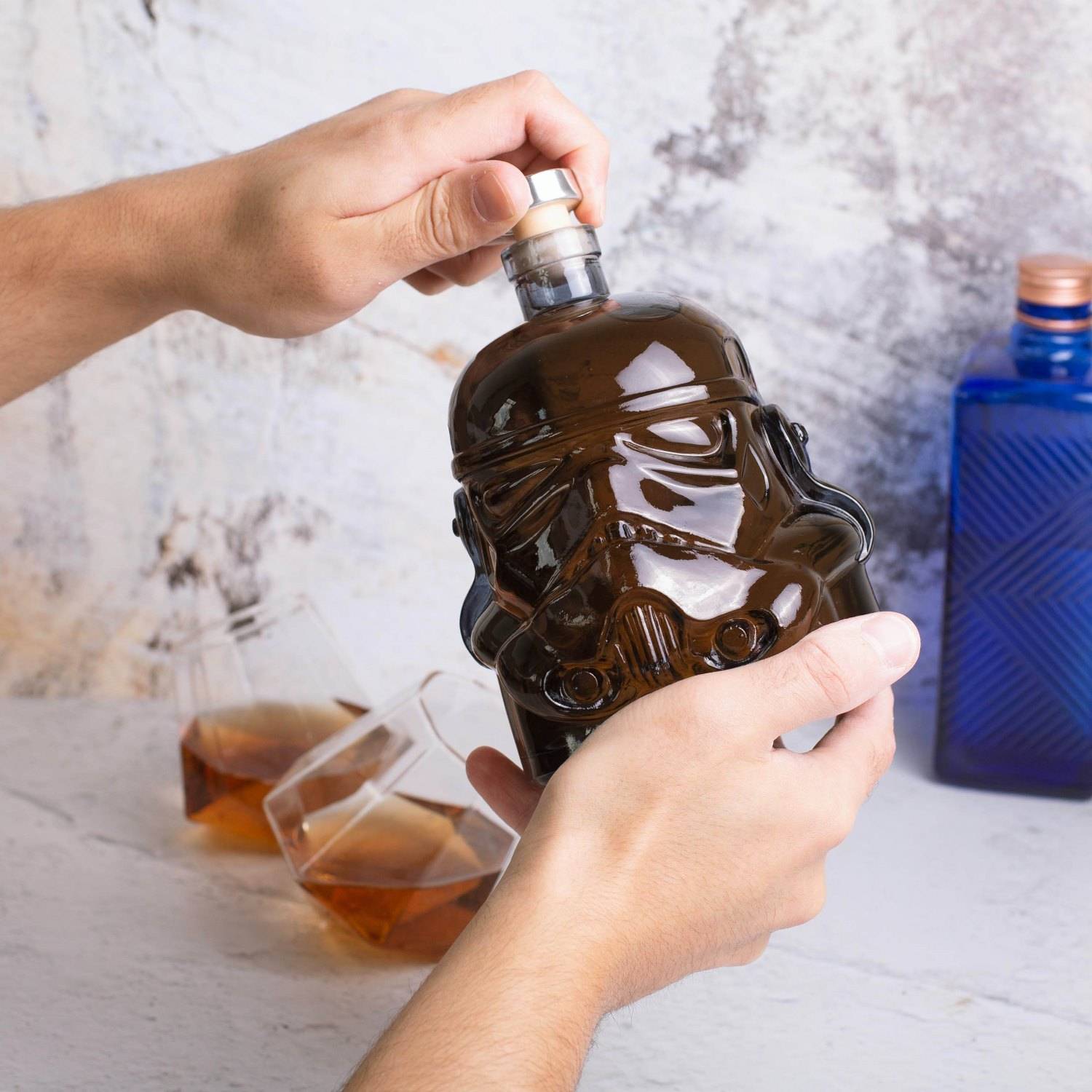 Hände halten eine schädelförmige braune Likörflasche, während sie diese auf einem Tisch mit zwei Gläsern und einer blauen Flasche im Hintergrund ausgießen oder öffnen.