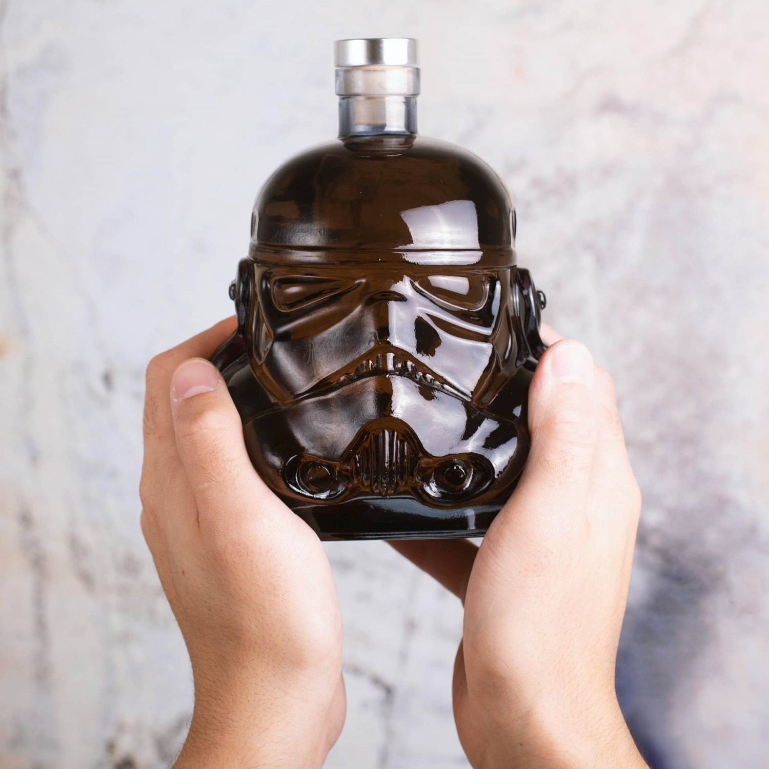 Hände, die eine dunkle Glasflasche in Form eines Stormtrooper-Helms auf hellem Hintergrund halten.